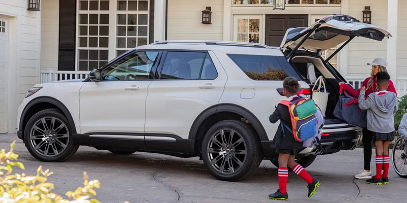 Two children prepare to load bags into the open trunk of a white SUV in a driveway.