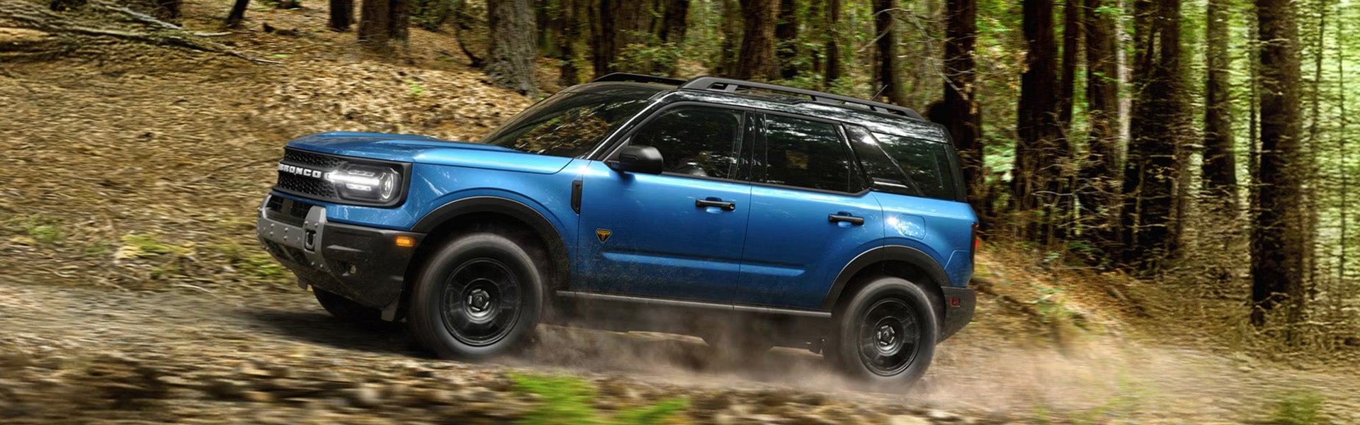 Blue Ford Bronco SUV driving on a dirt trail through a wooded forest.