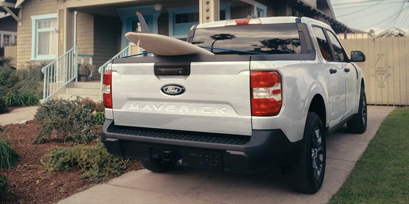A white Ford Maverick with a surfboard secured in the truck bed, parked against a sunny outdoor backdrop.
